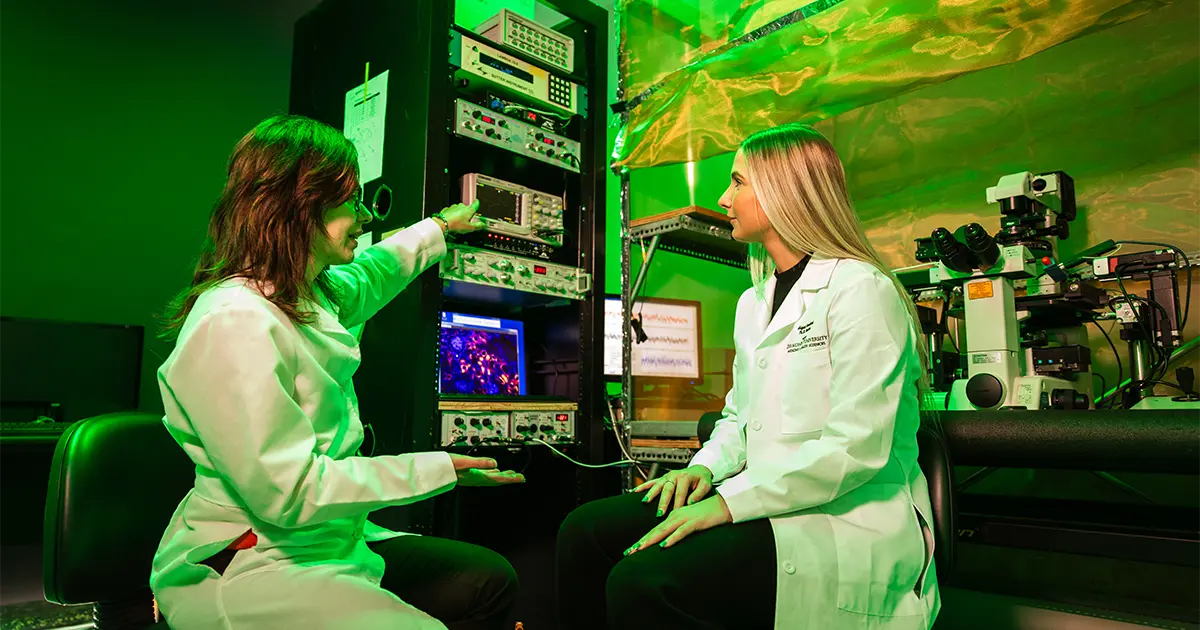 A DMU PhD student and faculty member in lab coats work together in a dimly lit research lab, reviewing data on specialized electrophysiology equipment beside a microscope.