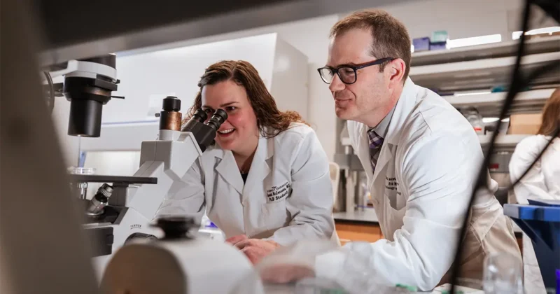 A DMU student and a faculty member in lab coats work together at a microscope, reviewing samples and discussing their findings in a research laboratory.