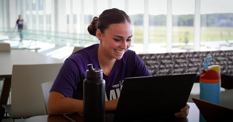 A student wearing a purple DMU shirt uses a laptop while smiling.