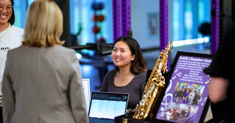 A DMU student speaks with visitors at a display table featuring a laptop and a music‑themed poster with a saxophone during the annual Club Day event.