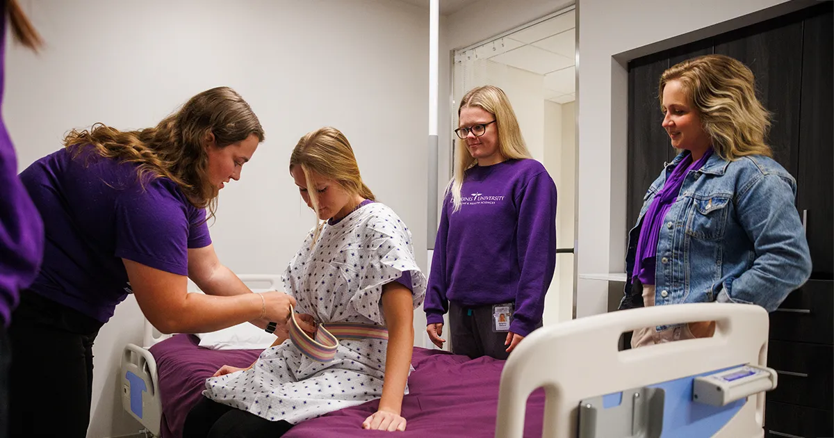 Occupational therapy students practicing home-care skills in a simulated bedroom environment with a client seated on a bed, adjusting adaptive equipment.