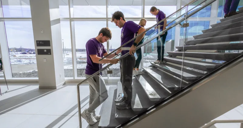 DMU students wearing purple shirts practice stair mobility techniques during a hands-on training exercise inside a modern campus building, while classmates observe from the staircase.