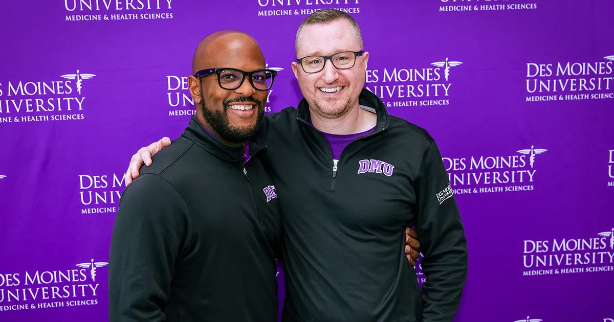 Two DMU employees pose for a photo in front of a purple backdrop.