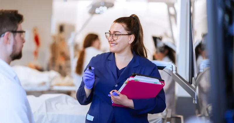 An instructor in a lab coat and gloves holds a marker and a stack of folders while speaking to a student in a clinical skills lab. Lab tables and equipment are visible in the background.