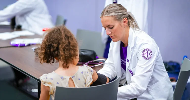 A DMU PA student in a white coat checks a young child’s blood pressure during the university’s annual Back to School Physicals event.