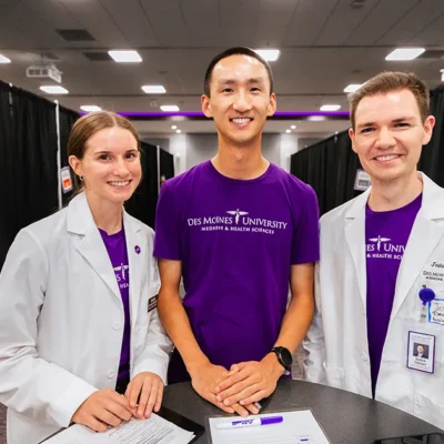 Three DMU students stand together at a registration table during the annual Back to School Physicals community event, wearing purple DMU shirts and white clinical coats in a large event space.