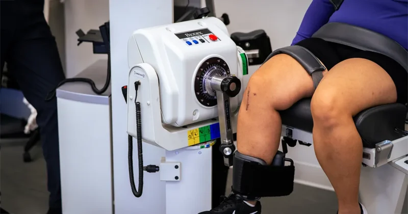 A person sits in a clinical testing chair with their leg secured in a Biodex isokinetic dynamometer. A visible surgical scar is present on their knee as the device measures strength and joint movement during testing.