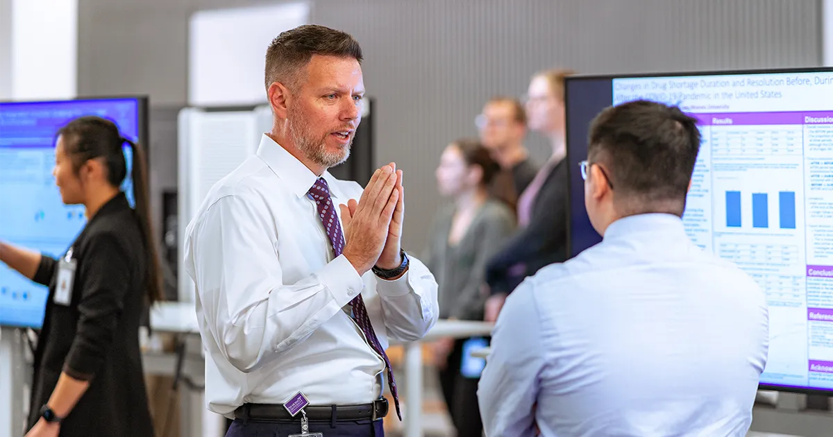 Two people stand in conversation during a research poster session, surrounded by presentation boards and attendees reviewing scientific charts and data.