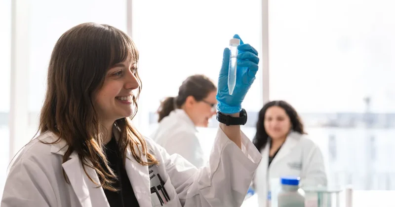 A DMU student wearing a lab coat and blue gloves examines a sample tube held up to the light while other students work in the background of a bright biomedical lab.