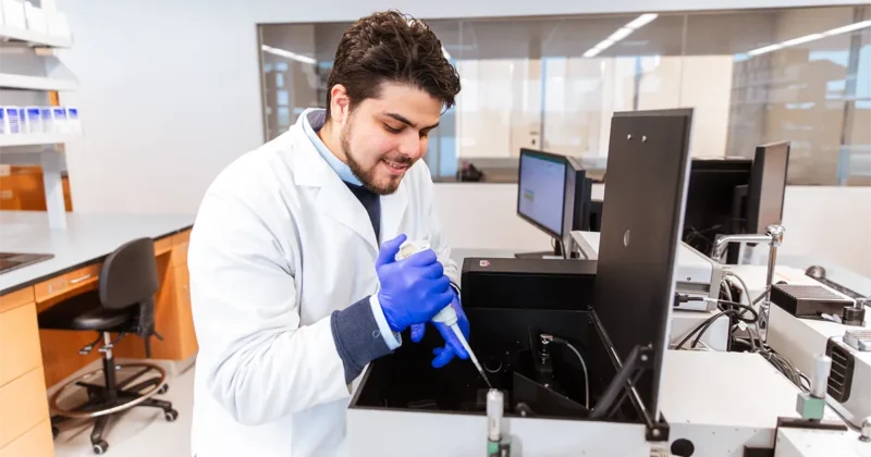 A DMU student wearing a lab coat and blue gloves uses a pipette while working with equipment in a biomedical research lab.