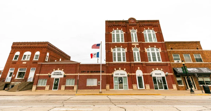 Street‑level view of the historic red‑brick municipal building complex in Manchester, Iowa, featuring the council chambers, city hall and police department. A flagpole with the U.S. flag and Iowa flag stand in front of the building.