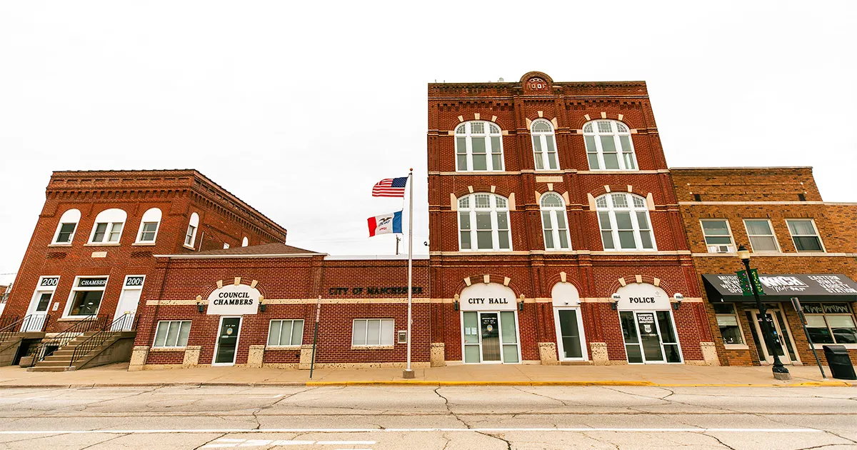 Street‑level view of the historic red‑brick municipal building complex in Manchester, Iowa, featuring the council chambers, city hall and police department. A flagpole with the U.S. flag and Iowa flag stand in front of the building.