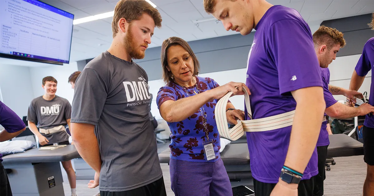A DPT faculty member demonstrates how to secure a gait belt around a student while other students observe and practice in a physical therapy lab.