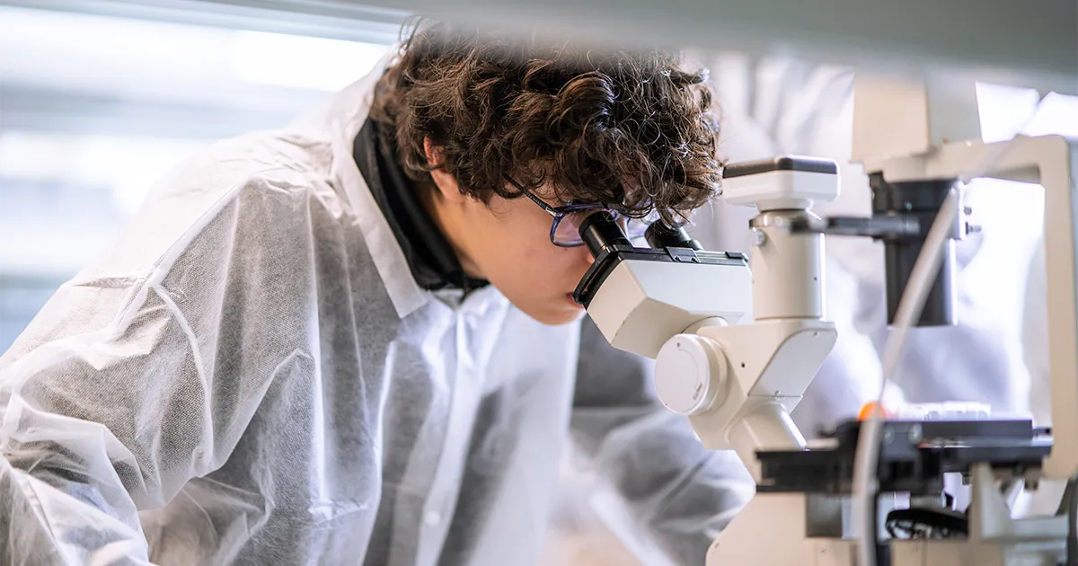 A student wearing a lab coat examines a specimen through a microscope during DMU’s Young Scientist Program.