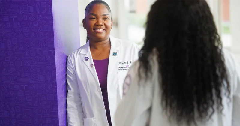 Two DMU students wearing white coats discuss course materials while leaning against a purple wall.