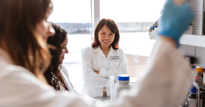 Two DMU students in lab coats work at a wet lab bench while a PhD program faculty member stands in the background observing the activity.