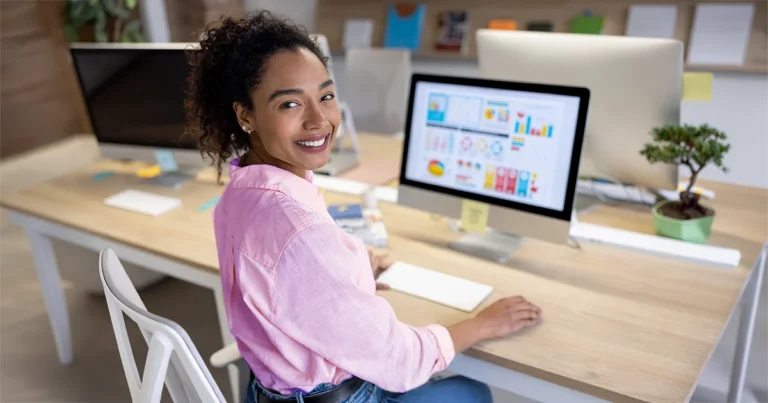 Person seated at a wooden desk in a modern office workspace, facing a desktop computer displaying charts, graphs and data visuals. The desk includes a keyboard, mouse, sticky notes and a small potted plant, with additional computer monitors and office supplies visible in the background.