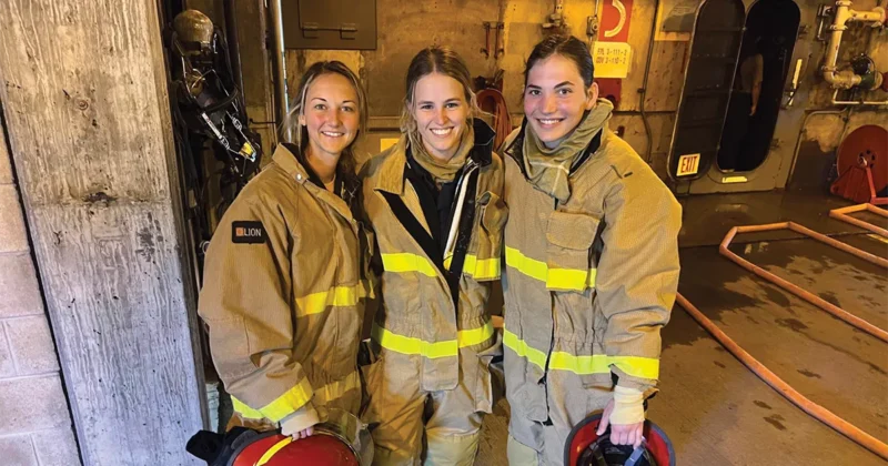 Three people wearing full firefighting gear stand together inside an industrial training area, each holding a helmet.