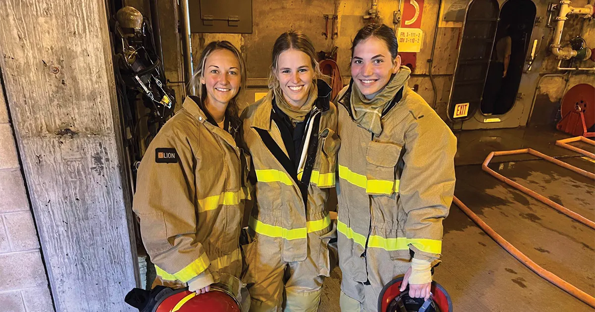 Three people wearing full firefighting gear stand together inside an industrial training area, each holding a helmet.
