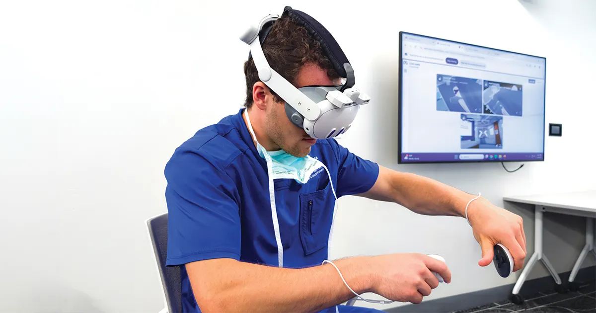 A DMU student wearing a virtual‑reality headset practices a hands‑on simulation using handheld controllers while a display screen shows related instructional content.