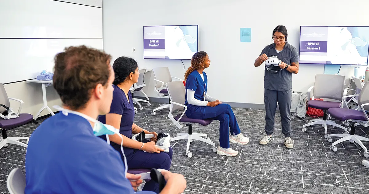 A DMU staff member leads a small group session on virtual‑reality equipment as other students watch and hold VR headsets in a classroom setting.