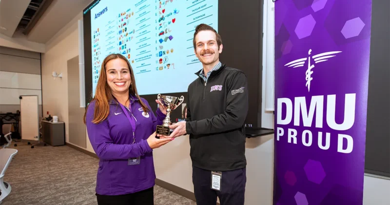 Jake Smith presents the Deans Cup to Jennifer Beaty, MD, during One Day for DMU. The scene is in a campus presentation space, with a screen and a DMU Proud banner in the background.
