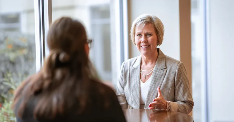 A DMU MHA alum speaks with another professional in a bright, modern office setting, gesturing as they discuss a topic across a table near large windows.