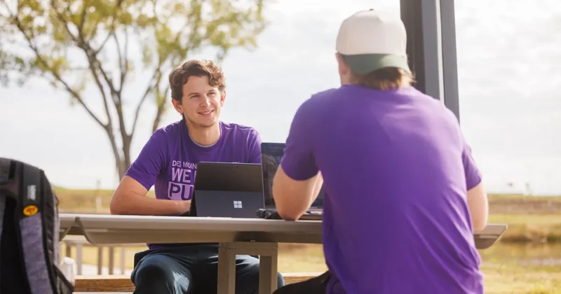 Two DMU students sit at a table outside near the on-campus pond.