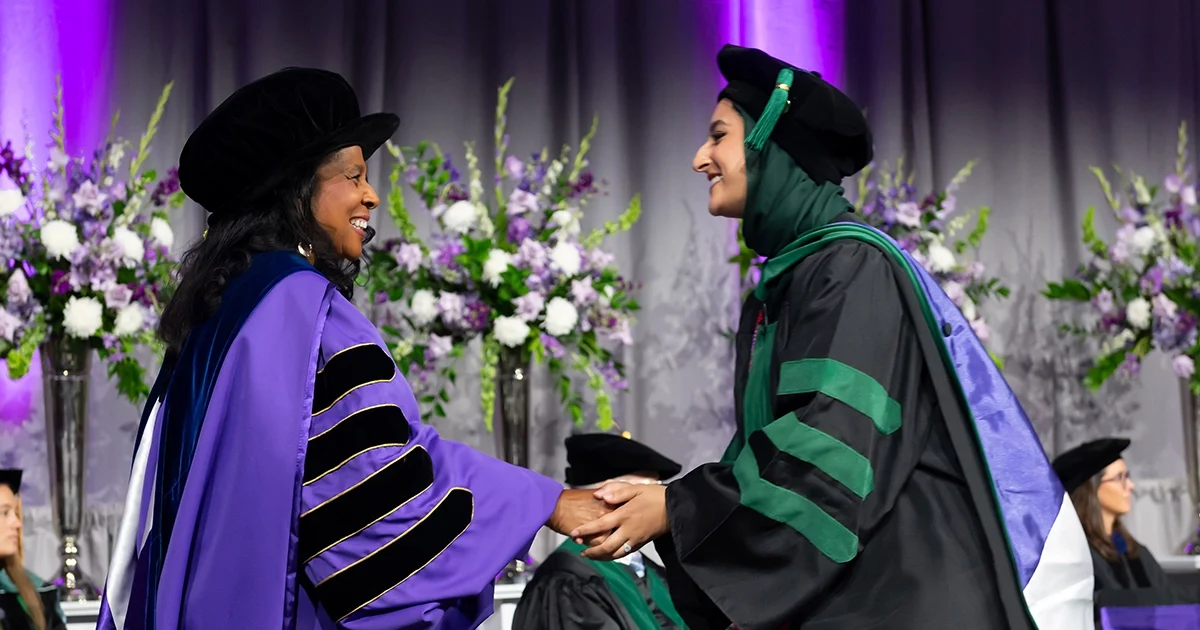 DMU president and CEO Angela L. Walker Franklin at left, in purple academic regalia, shakes hands with a graduating DMU student in green- and black-trimmed academic gown and cap on a commencement stage, with large floral arrangements and purple lighting in the background.