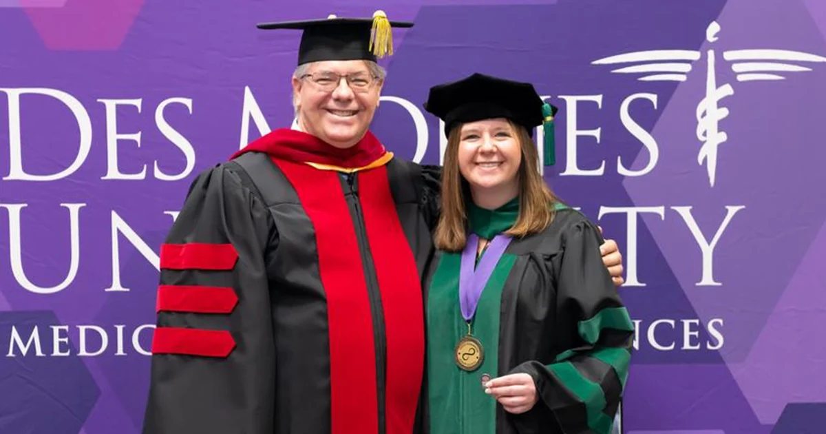 Two DMU Grand Family members in academic regalia pose together in front of a purple DMU backdrop; one wears a black gown with red trim and cap, and the other is a DMU student in a green- and black-trimmed gown and cap, holding a medal on a ribbon.