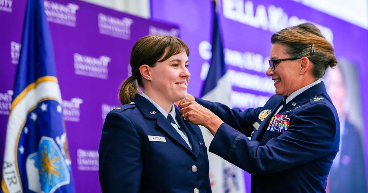 A DMU student in a U.S. Air Force dress uniform stands at the front of a stage while another uniformed individual pins an insignia to the student’s collar during a commissioning ceremony, with a purple DMU-branded backdrop and flags in the background.
