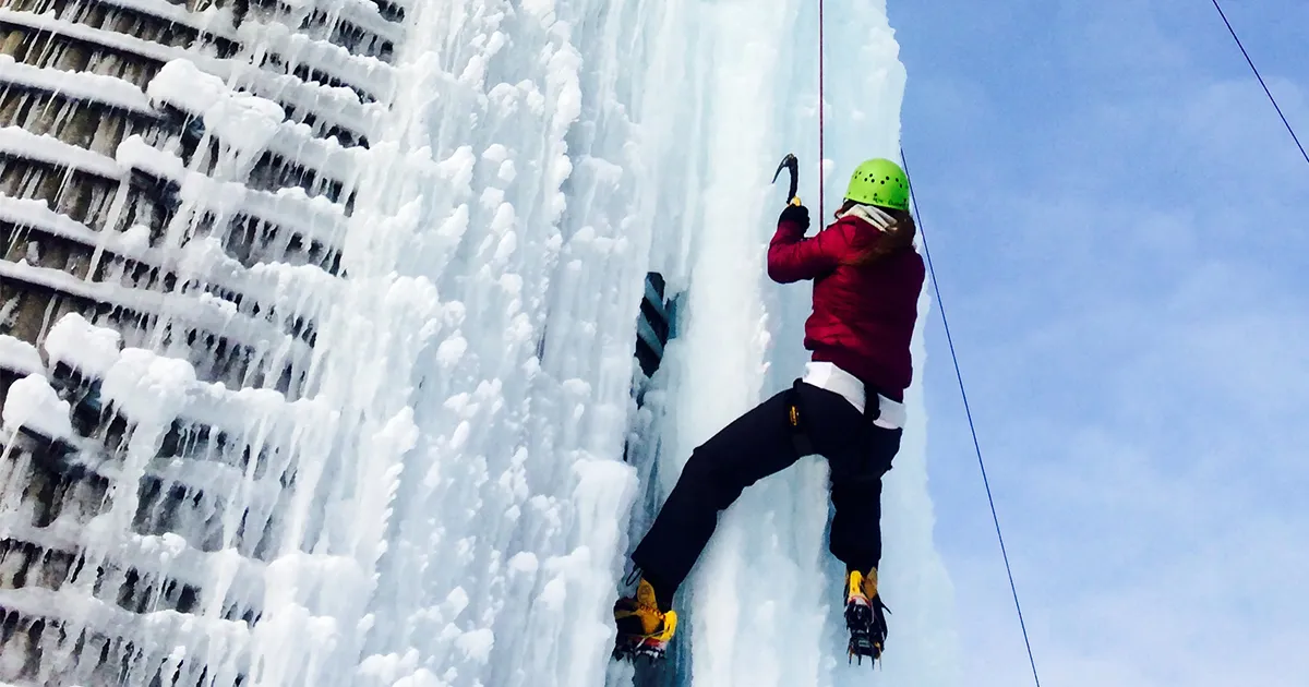 Libby Abbas, DO’15, wears a helmet, harness and crampons while climbing a frozen ice wall using an ice axe, secured by a rope against a winter sky.