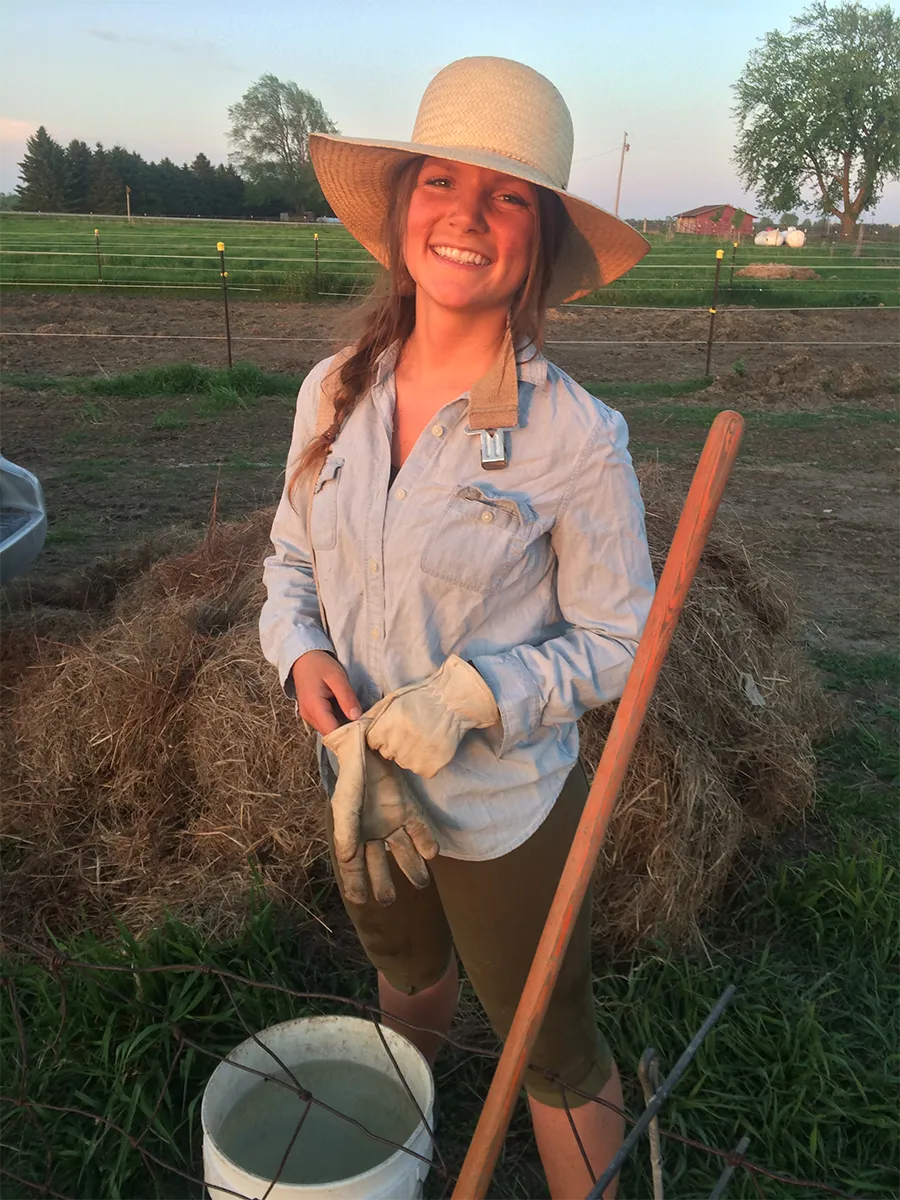 Libby Abbas, DO’15, in a wide-brimmed hat, work gloves and a long-sleeved shirt stands beside a hay bale in a field, holding gloves near a bucket and long-handled tool at sunset.
