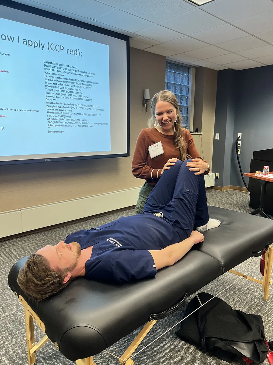 Libby Abbas, DO’15, stands beside a treatment table and performs a hands-on musculoskeletal assessment on an individual lying on the table, with a presentation slide projected on a screen in the background.