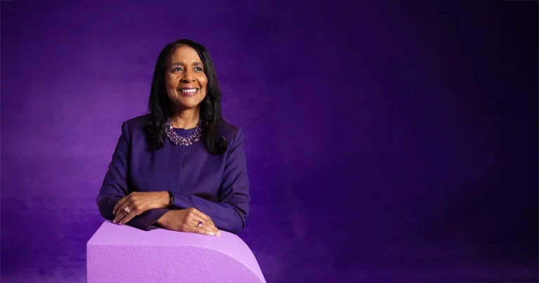 Angela L. Walker Franklin, Ph.D., poses for a portrait while resting her arms on a large letter "D." Franklin wears a purple suit and necklace, standing against a dark purple studio background.