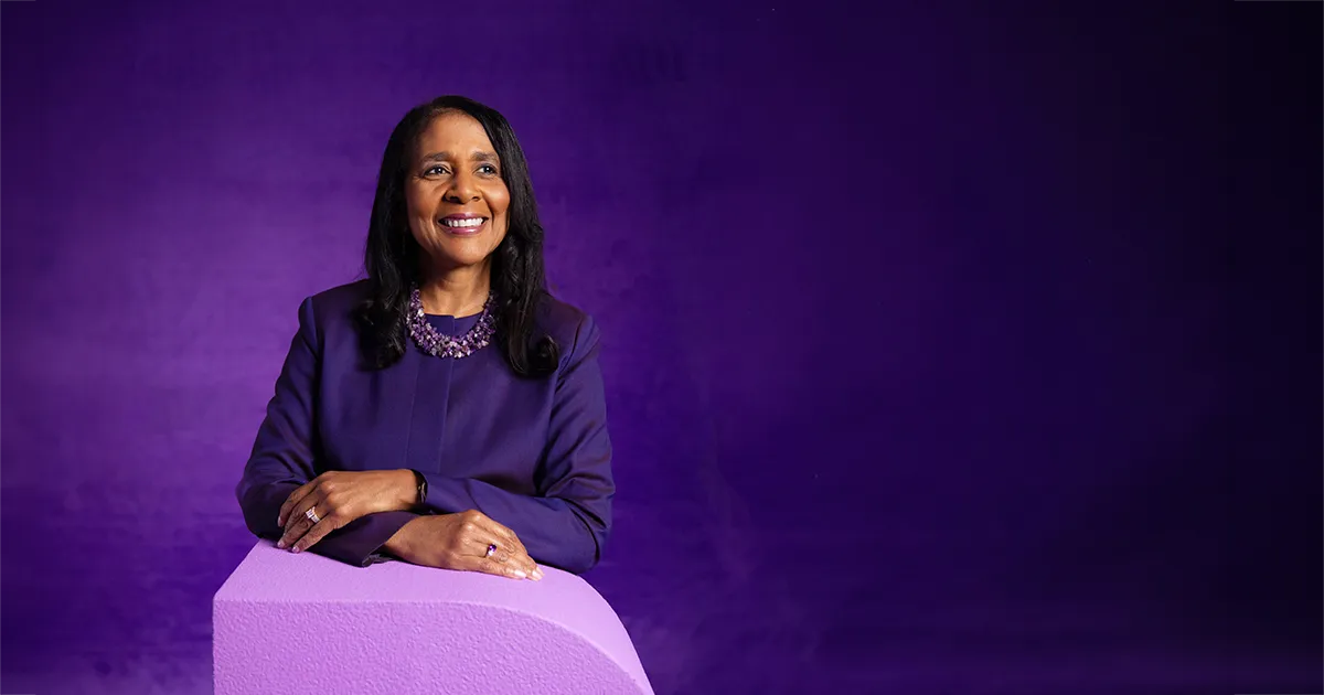 Angela L. Walker Franklin, Ph.D., poses for a portrait while resting her arms on a large letter "D." Franklin wears a purple suit and necklace, standing against a dark purple studio background.
