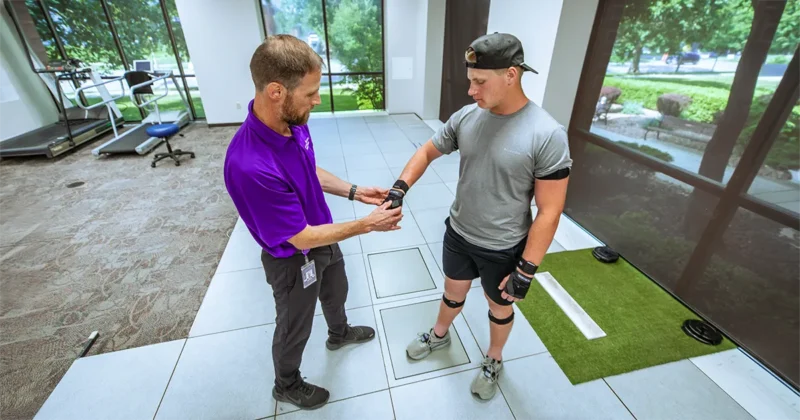 Shane McClinton adjusts wearable motion-tracking sensors on a participant standing on a motion-analysis platform in DMU’s Human Performance Lab at DMU 32. Exercise equipment and large windows overlooking a landscaped outdoor area are visible in the background.