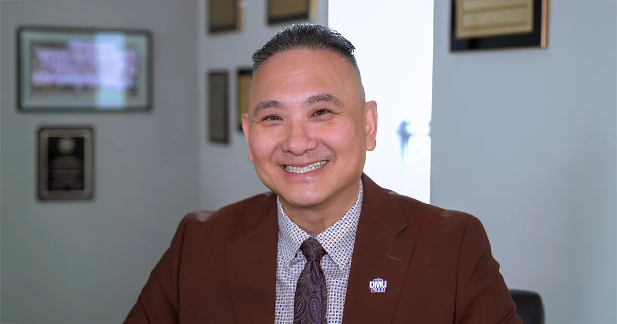 John T. Pham, DO’00, FACOFP, seated in an office wearing a brown suit jacket with a DMU Proud lapel pin.