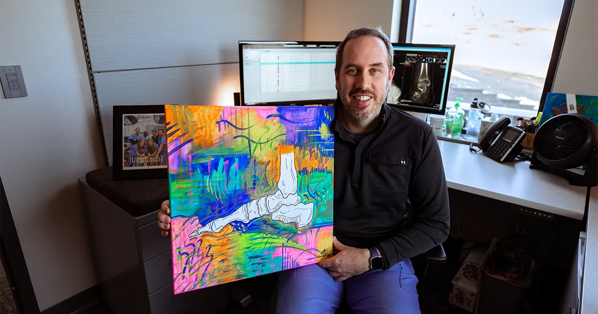 Jeffrey Dikis, DPM’12, sits in a medical office holding a colorful painting of a foot anatomy illustration, with computer monitors in the background.
