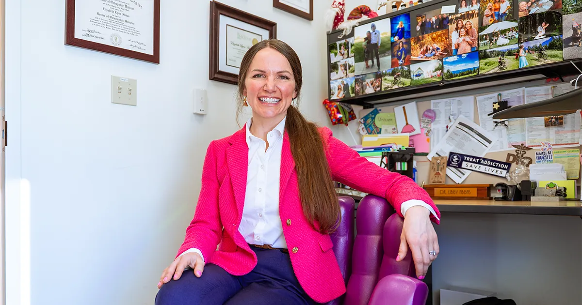 Libby Abbas, DO’15, sits in a purple chair in an office, wearing a bright pink blazer over a white shirt. Framed diplomas hang on the wall behind her, and a bulletin board displaying family photos and mementos is visible above a desk.