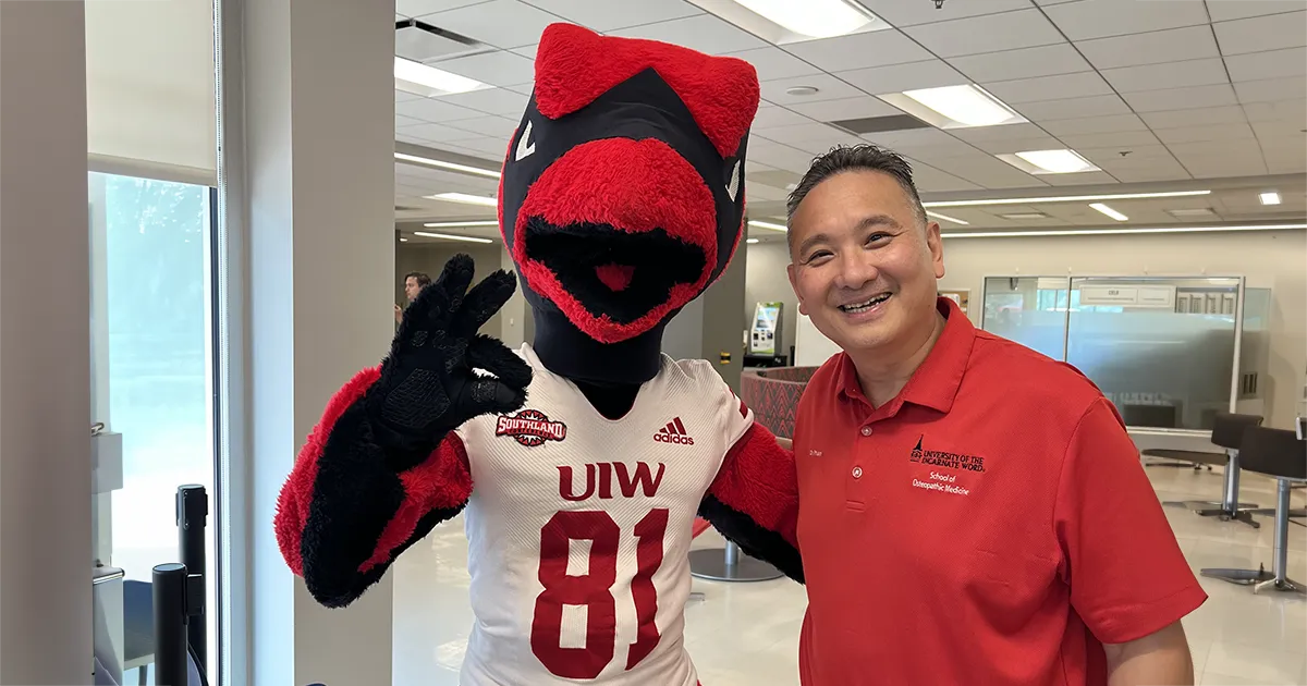 John T. Pham, DO’00, FACOFP, stands beside the University of the Incarnate Word Cardinals mascot inside a campus building.