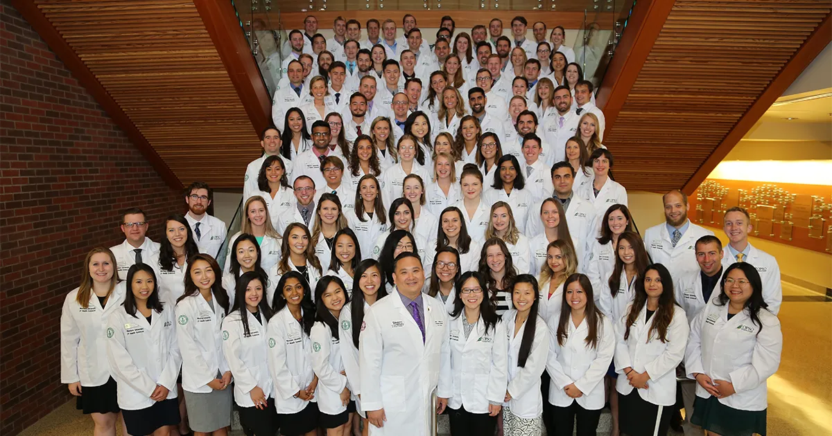 John T. Pham, DO’00, FACOFP, poses with a large group of medical students wearing white coats on a staircase.