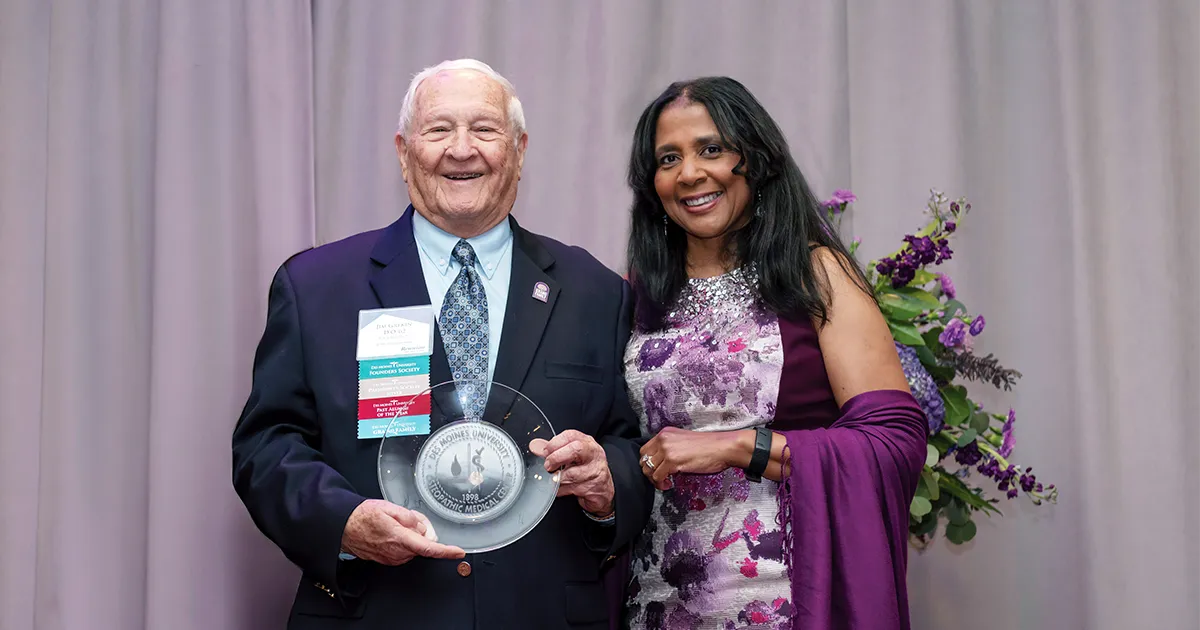 President Angela L. Walker Franklin, PhD, poses with James A. Grekin, DO’62, MACOI, as Grekin holds a commemorative award during a recognition event. A floral arrangement and draped backdrop are visible behind them.