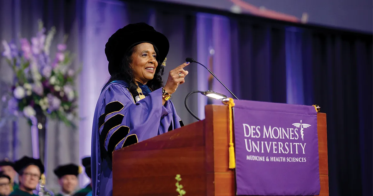 President Angela L. Walker Franklin, PhD, delivers remarks from the podium during a DMU Commencement ceremony. Wearing academic regalia, she addresses the audience from a stage decorated in DMU colors.