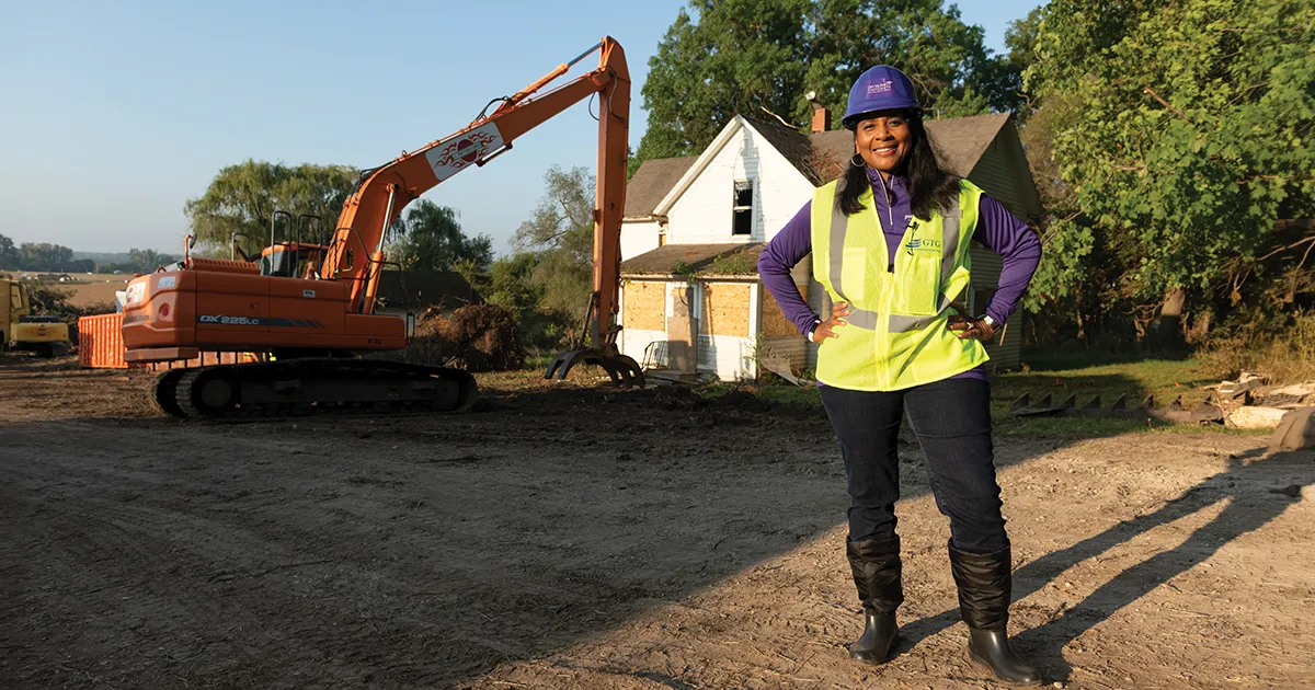 President Angela L. Walker Franklin, PhD, stands at a construction site wearing a hard hat and safety vest as an excavator clears land beside a farmhouse slated for demolition at 8025 Grand Ave.