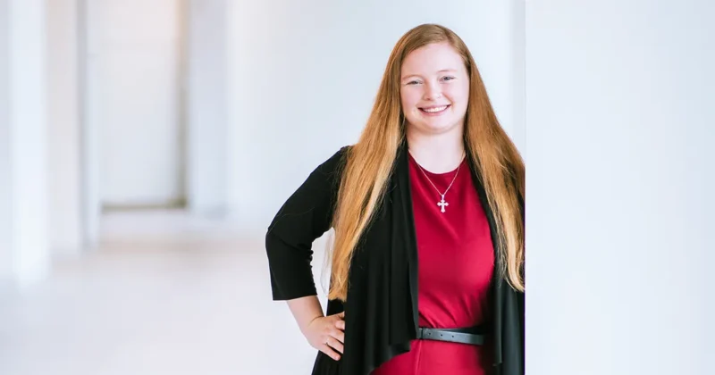 Kassidy Hackbarth, OTD’26, stands in a campus hallway wearing a red outfit and black cardigan