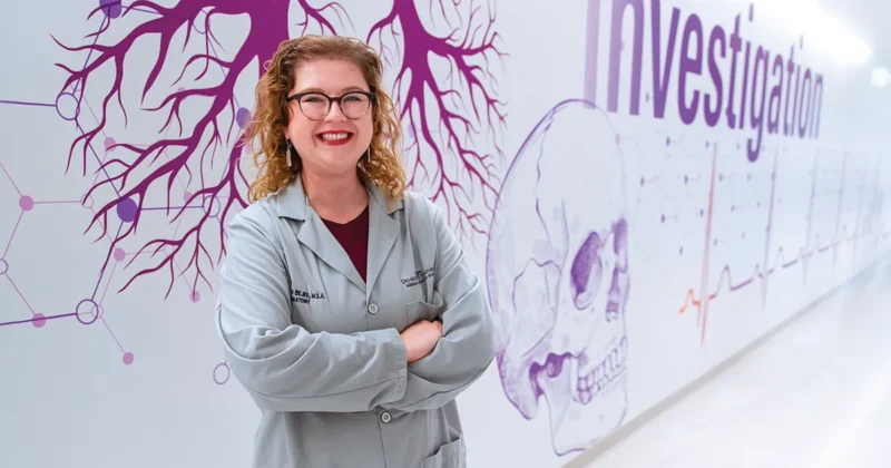 Copilot said: Katie Bejes, MSA’20, stands in a DMU hallway wearing a lab coat in front of a wall mural featuring anatomical illustrations, vascular structures and the word “Investigation.”