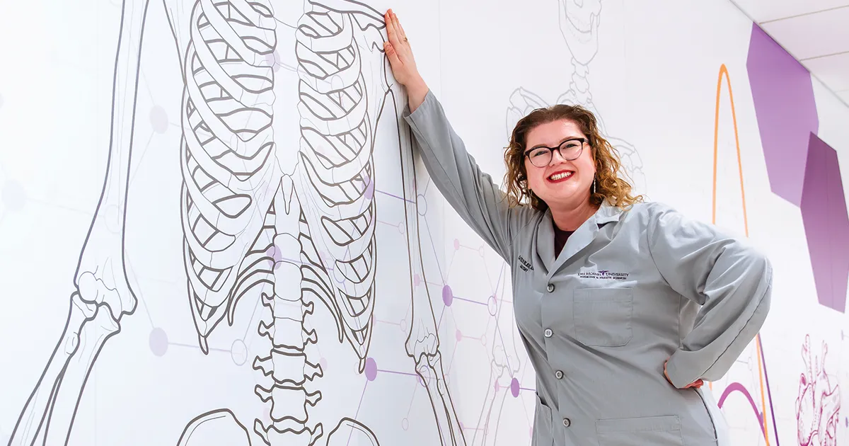 Katie Bejes, MSA’20, stands in a lab coat beside a large anatomical skeleton illustration displayed on a wall featuring medical and scientific graphic elements.
