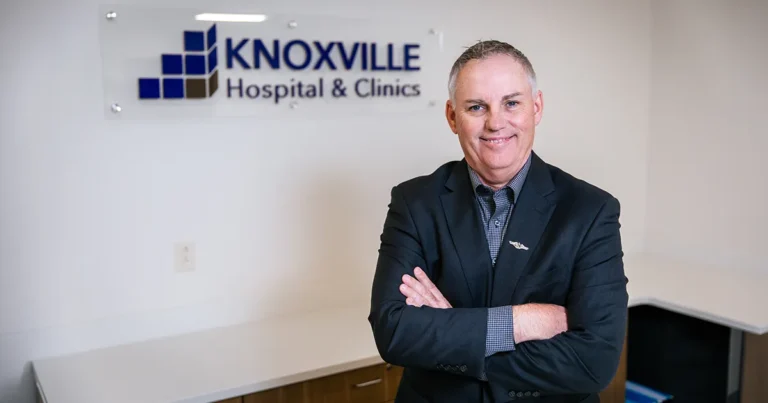 Kevin Kincaid, MHA’09, stands with arms crossed in an office at Knoxville Hospital & Clinics with a hospital logo displayed on the wall behind him.