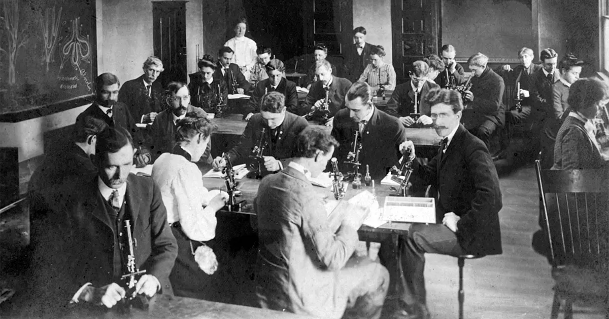 Historic photograph of students working at microscopes in a laboratory classroom in 1905, seated at long tables with instructors and laboratory equipment throughout the room.
